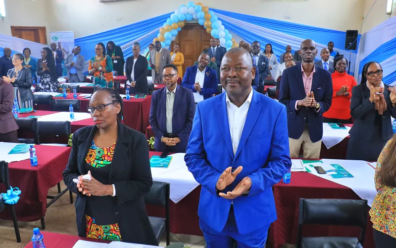 A diverse group of people stands applauding in a decorated hall with blue and gold balloons.