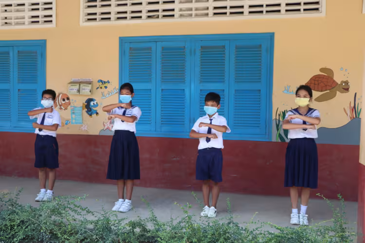 Four students in uniforms and masks standing outside and creating an equal sign with their arms. 