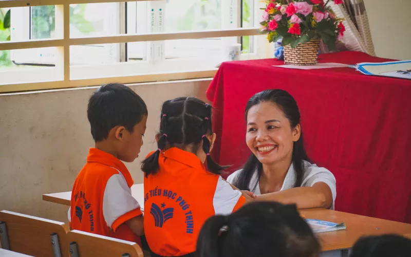 A teacher smiles while interacting with two children in orange uniforms at a school desk.