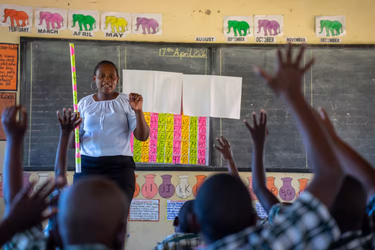 A teacher in a Zambian classroom stands in front of a chalkboard, while students raise their hands to answer.