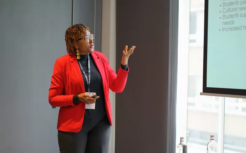 Cleopatra is in red blazer and presents confidently beside a screen displaying bullet points. Her expression is engaged, creating an informative atmosphere.