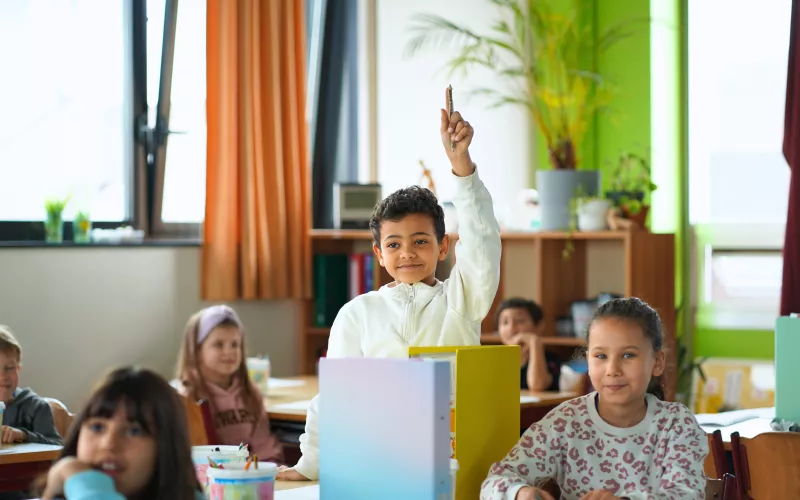 A young boy in a classroom eagerly raises his hand, surrounded by attentive classmates. The room is bright, with plants and colorful dividers.