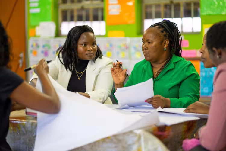 Female eachers sit together at a classroom table discussing notes during a professional learning community.