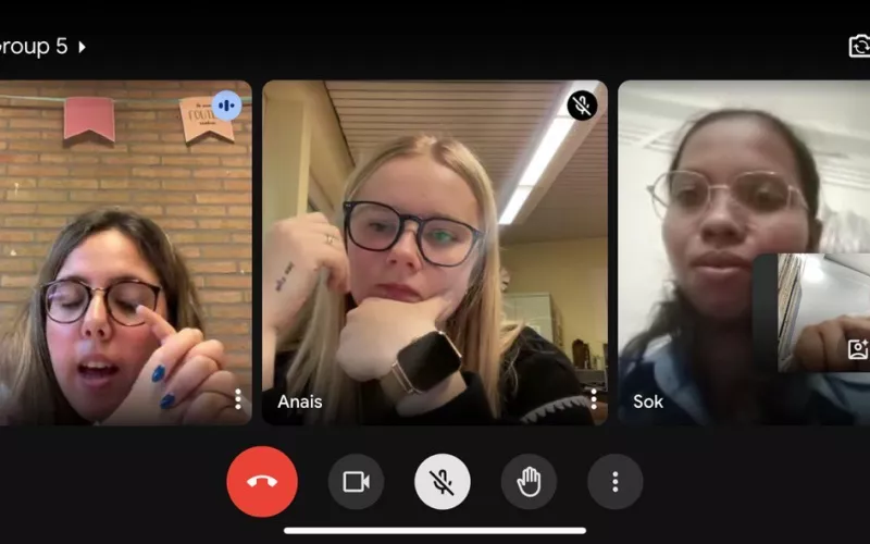 A video call with three people, each in their own video window. The mood appears focused. One person has a background with brick and banners.