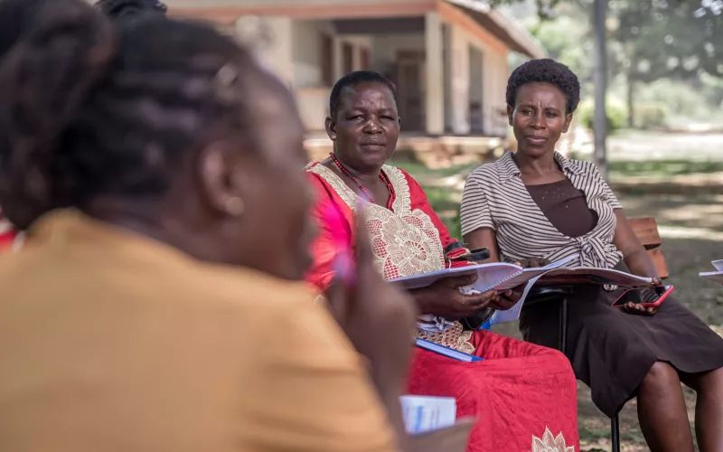 Female teachers sit outdoors with notebooks, listening during a group discussion.