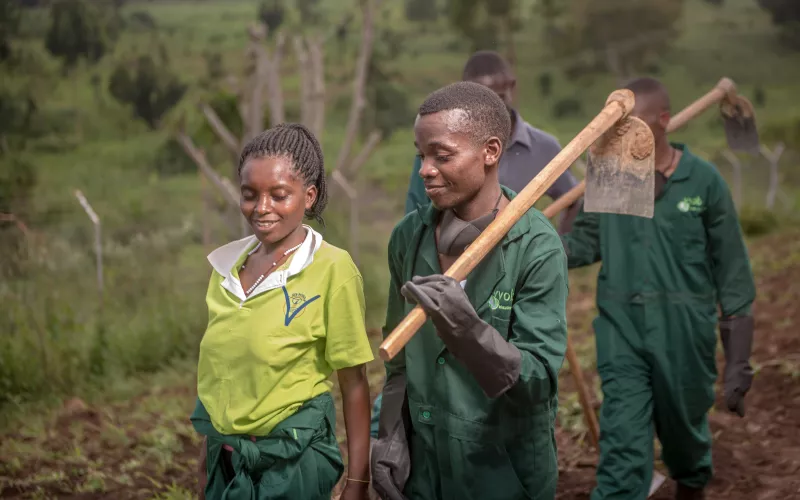 Students in green overalls carry hoes while walking together on farmland.