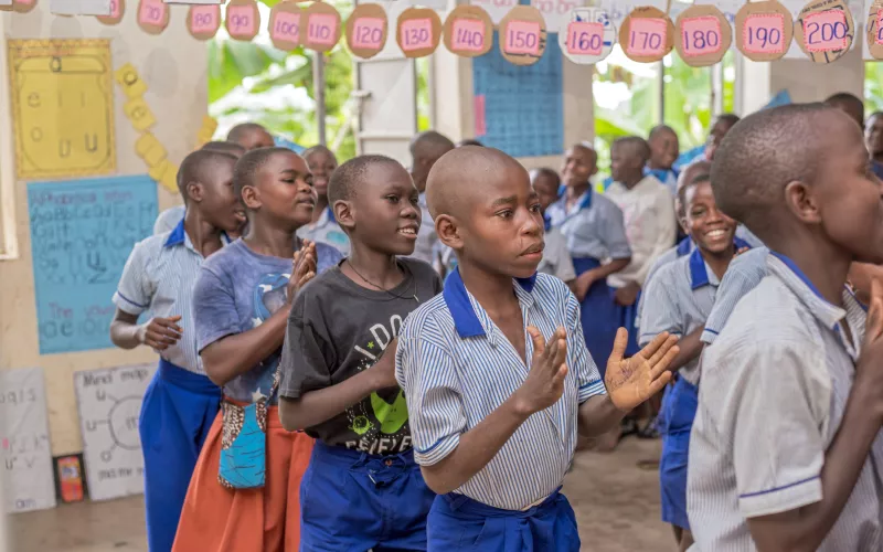 Children in blue uniforms and one in a black t-shirt are clapping in a colorful classroom, decorated with educational posters and paper numbers. They appear joyful and engaged.