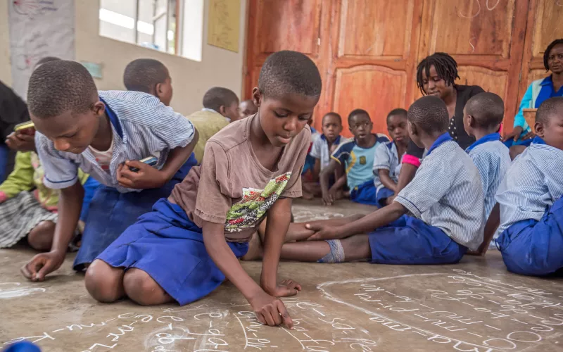 Young learners write on the classroom floor with chalk during a group literacy activity.