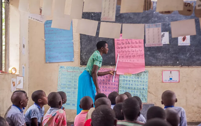 A teacher stands at the front of a classroom pointing to a pink chart with letters, while young students sit on the floor attentively watching.
