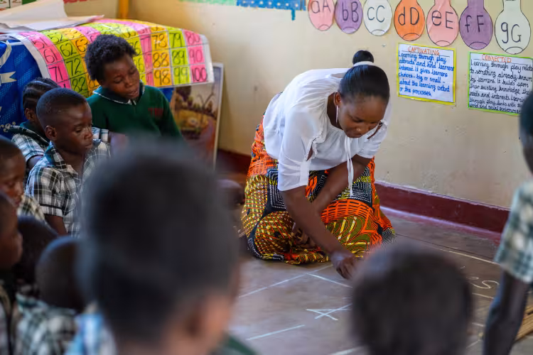 A teacher in a Zambian classroom writes on the floor with chalk, surrounded by learners.
