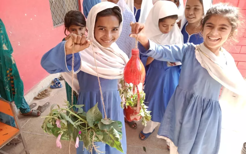 Smiling girls in blue and white uniforms hold hanging plant crafts made from recycled bottles. They stand outside a red wall, exuding happiness and creativity.