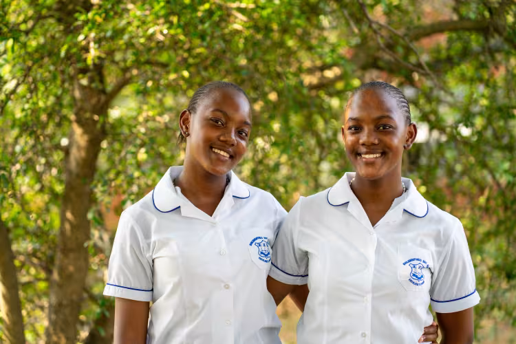 Two smiling twin sisters in white school uniforms with blue trim, standing closely together in front of a lush, green, leafy background.