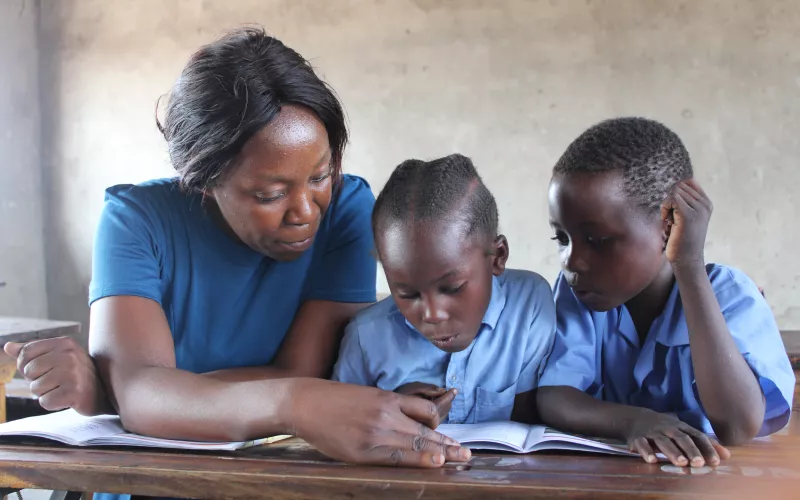 A teacher sits with two children in uniforms at a desk, pointing at a book. The children focus intently, creating a scene of learning and guidance.
