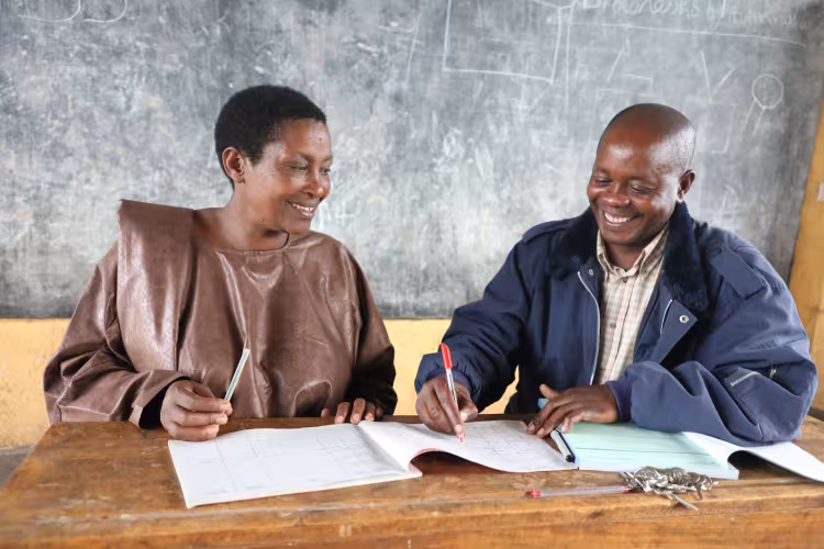 Two people sit at a wooden desk, smiling as they write in notebooks. Behind them is a chalkboard with faint diagrams. The atmosphere is warm and collaborative.