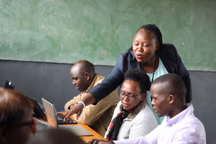 A teacher assists three adults with laptops in a classroom with a green chalkboard. She gestures energetically, fostering a focused and collaborative atmosphere.