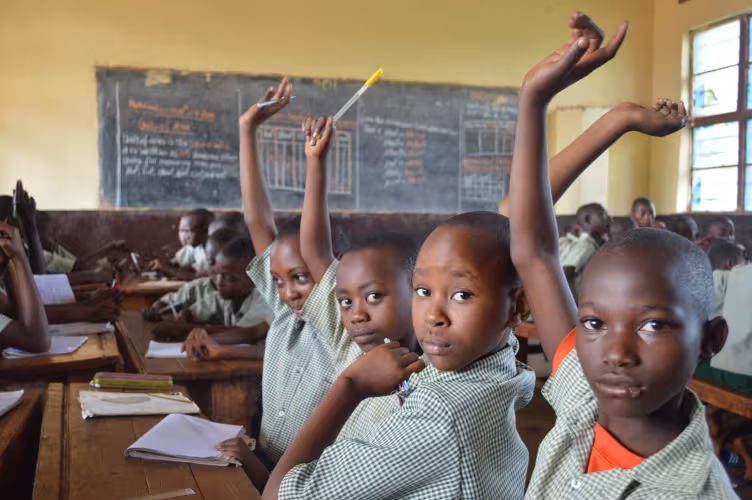 In a classroom, several students in uniforms eagerly raise their hands, signaling engagement. A chalkboard with writing is visible in the background.