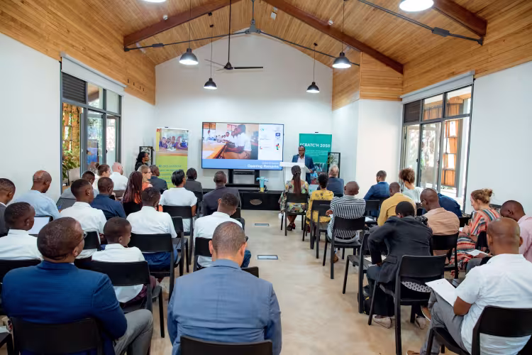 A group of people seated in a bright, modern conference room facing a large screen displaying a presentation. The room has wooden ceilings and large windows, creating an engaging and focused atmosphere.