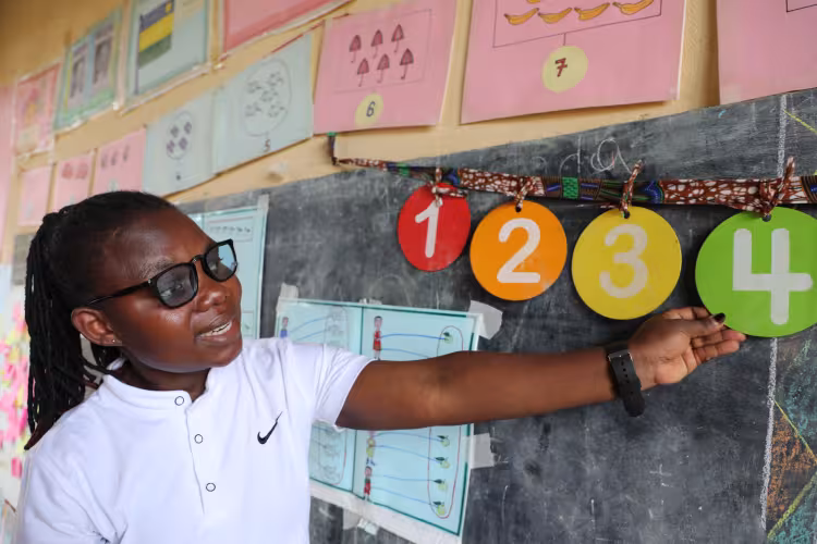 Female teacher in a classroom pointing to colorful number cards 1, 2, 3, 4 on a chalkboard, teaching counting skills with educational posters and math visuals in the background.