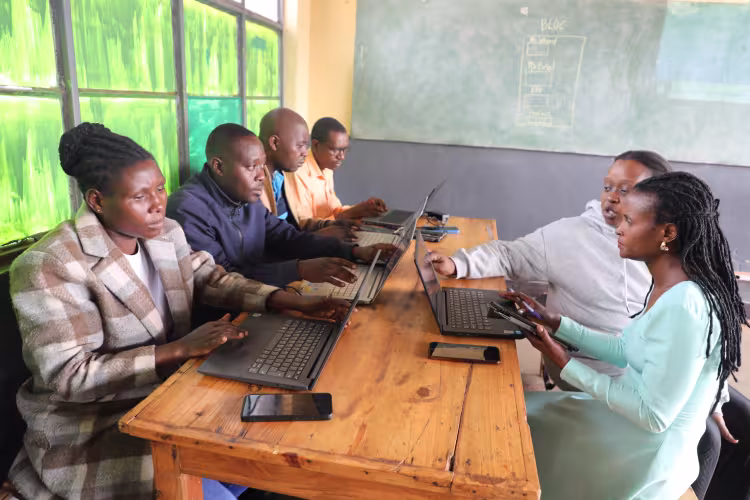 Five African professionals collaborating at a wooden table with laptops and smartphones in a classroom setting, participating in a digital skills workshop or team meeting. A green chalkboard behind them displays the word “BLOG” with a simple content outline diagram, highlighting blogging, content creation, and technology training.