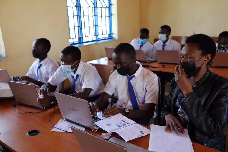 Students and a teacher, wearing masks, are engaged in a classroom, using laptops at wooden desks. The atmosphere is focused and collaborative.