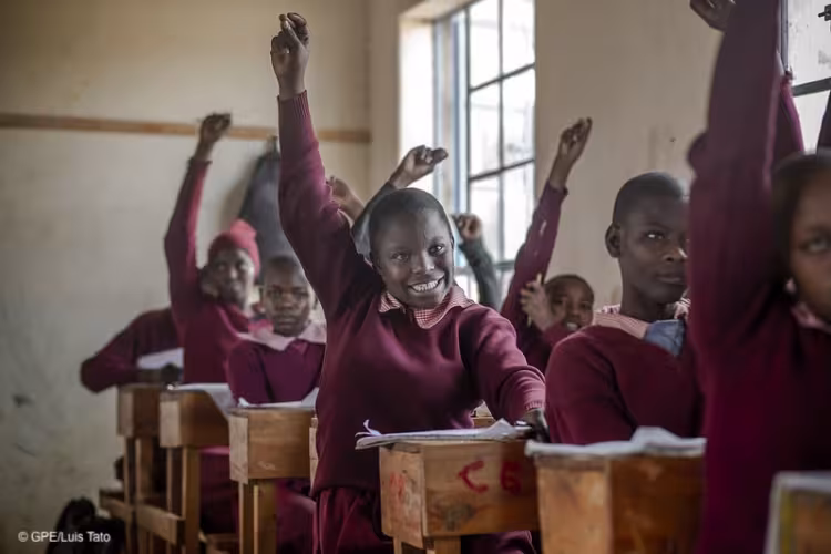 Primary school students in maroon uniforms seated at wooden desks, raising their hands eagerly to answer a question in a classroom.