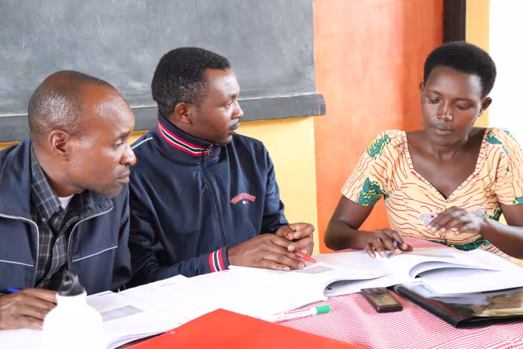 Three adults seated at a table reviewing documents together, engaged in discussion and collaborative study, with open books and notebooks in a classroom setting.