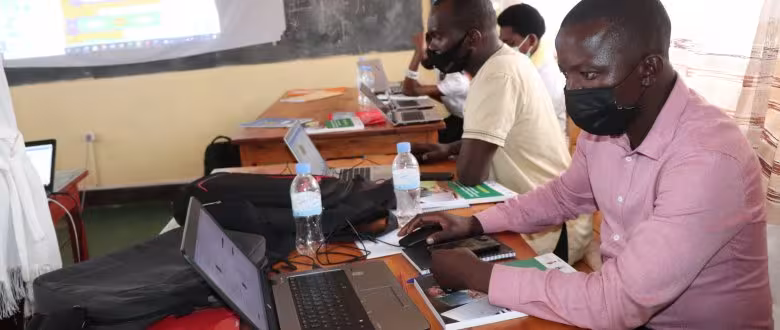 A group of people sit in a classroom setting, using laptops. Each person wears a mask, focused on their screens. Bottles of water and notebooks are on the desks, creating a studious atmosphere.