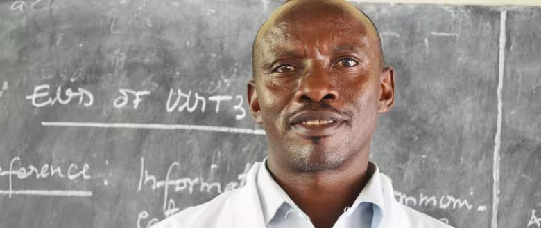 A man in a white coat stands in front of a chalkboard with handwritten notes. He looks confident and engaged, suggesting an educational setting.