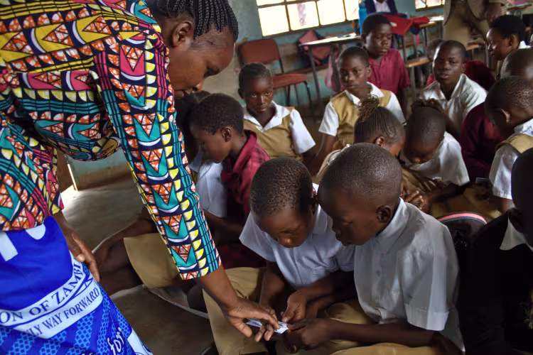 A teacher helps students with an activity in a classroom, where children in uniforms are seated closely together, focused on their work.
