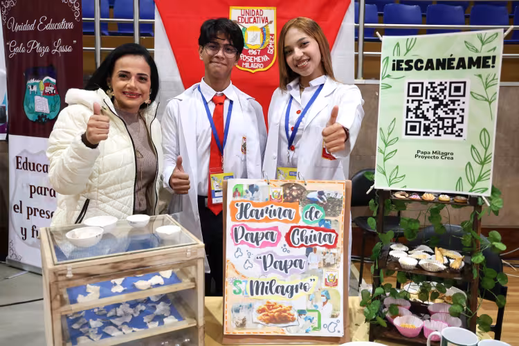 Three people pose with thumbs up at an educational fair booth. Two students in white lab coats stand beside a woman in a white jacket, surrounded by displays about making flour and food products from Chinese potato, including samples, a drying box, school banners, and a large QR code sign.