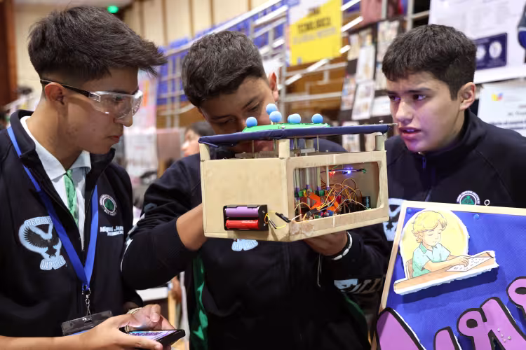 Three students examine a handmade electronic prototype at an educational fair. The wooden model has visible wiring, batteries, lights, and mechanical parts, while one student holds a smartphone beside a display about Braille.