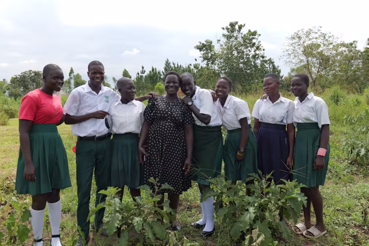 Smiling teacher and seven schoolgirls pose in a rural garden, wearing uniforms and standing among green plants under a bright sky.