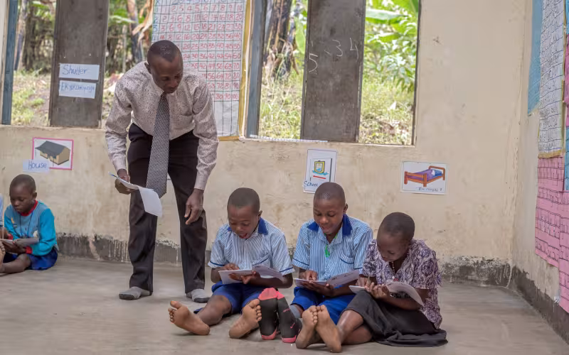 A teacher bends to guide three schoolchildren writing in notebooks on the classroom floor; another child sits nearby in a focused, calm lesson.