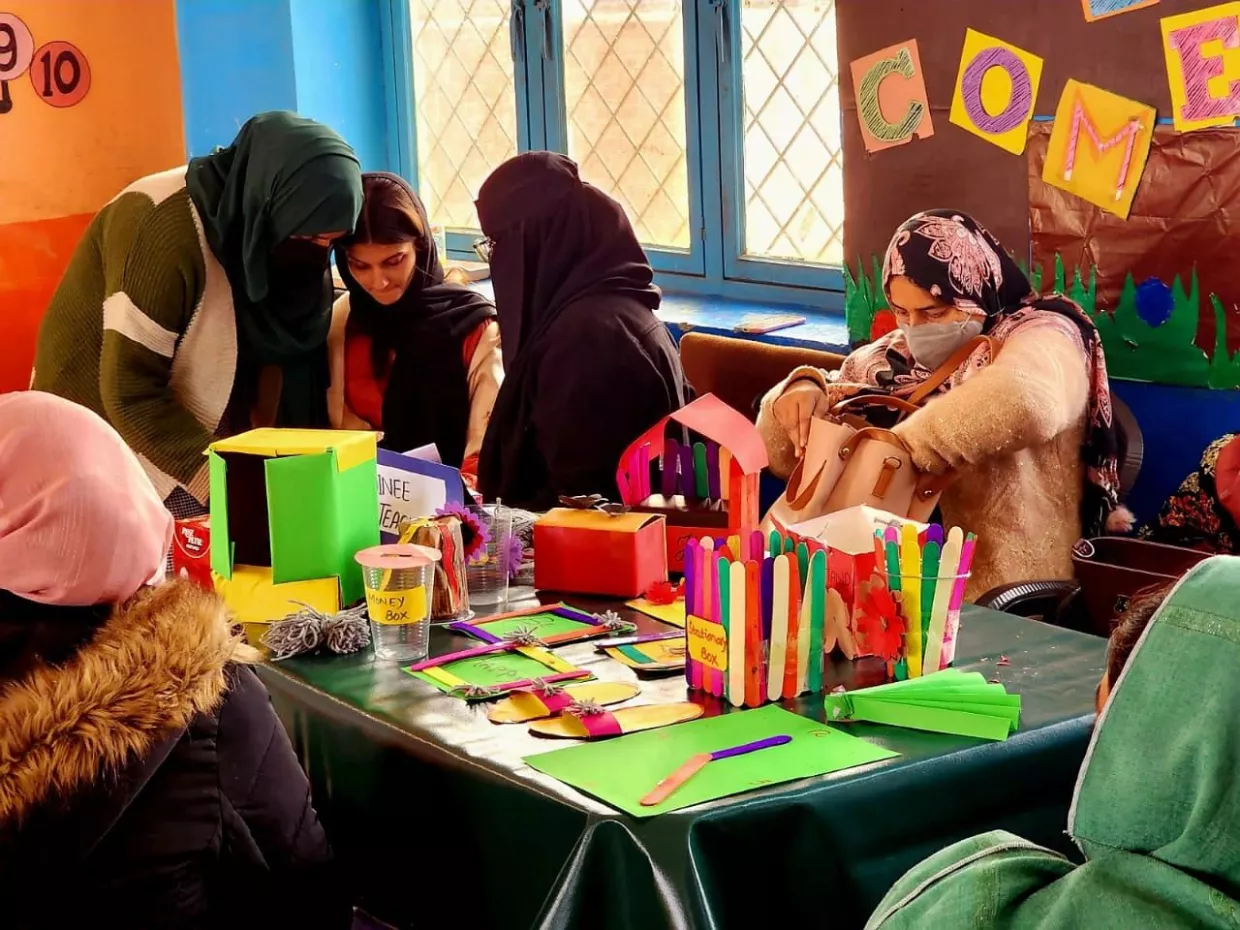 A group of women wearing headscarves and masks sits at a table with colorful craft materials, like popsicle sticks and paper, in a warm, welcoming room.