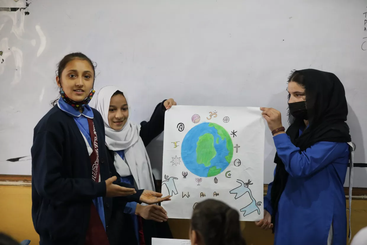 Three students present a colorful drawing of Earth with symbols and animals, smiling and engaged, in a classroom setting with a warm, collaborative tone.
