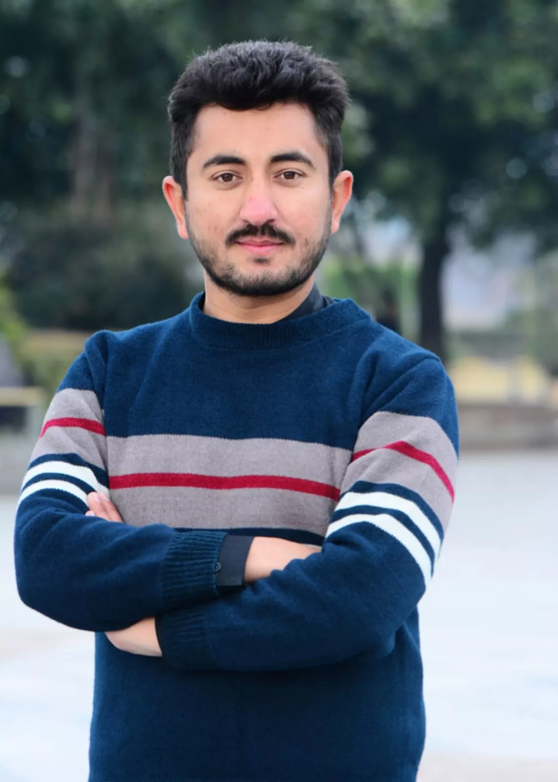 A man with short dark hair and a beard stands outdoors, arms crossed. He wears a blue sweater with red and white stripes. The background is blurred greenery. The expression is confident and relaxed.