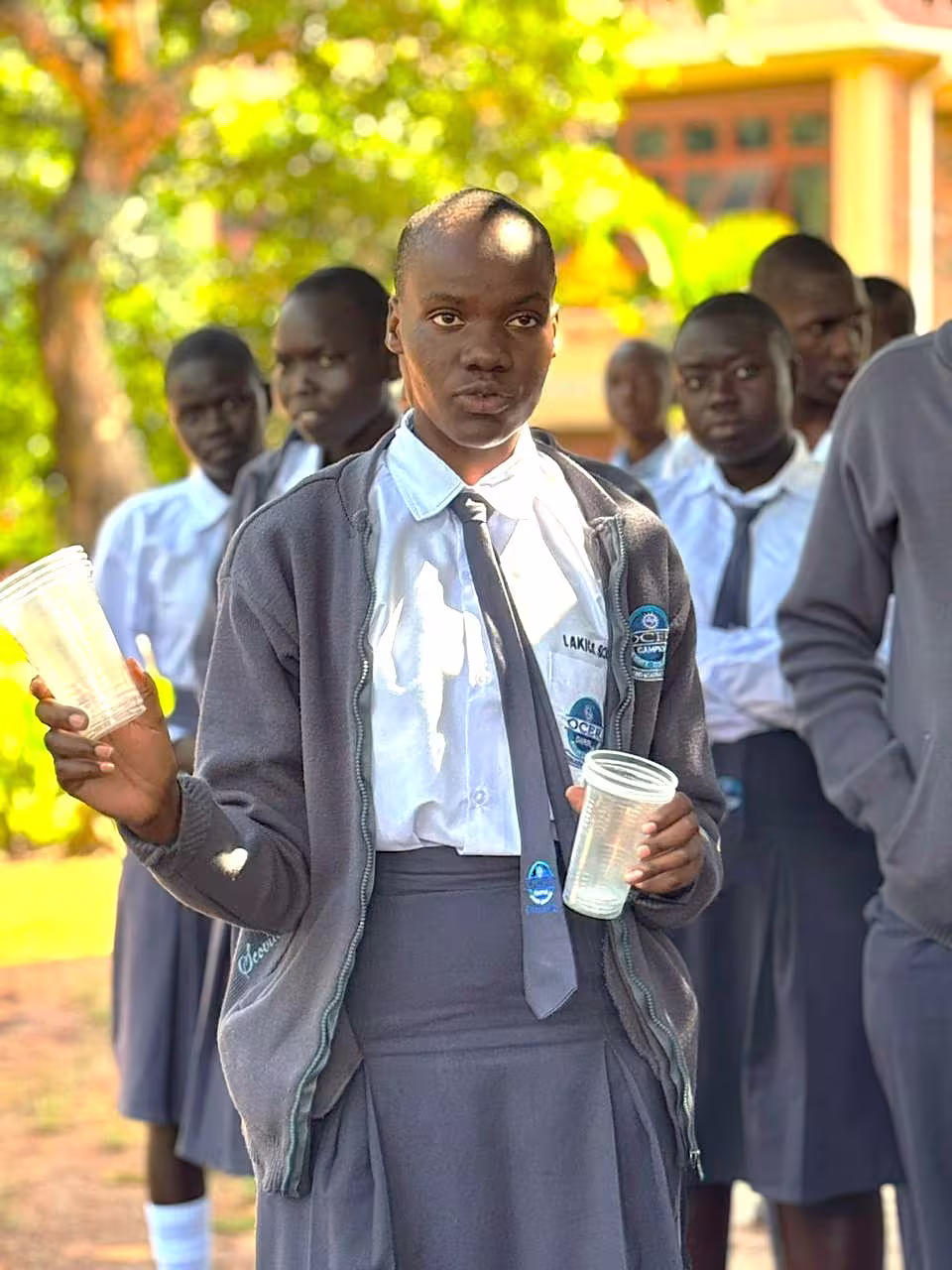 A student wearing school uniform and holding plastic cups during an outdoor activity while other students look on.