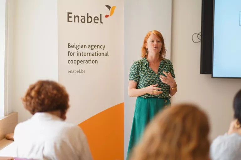 Woman in a green dress speaks passionately at a seminar, next to an Enabel banner, with seated audience members in the foreground.