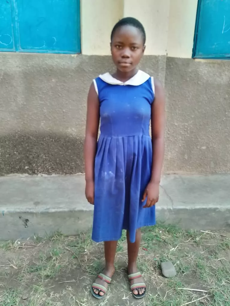 Esther is standing outside a school building wearing a blue uniform dress.