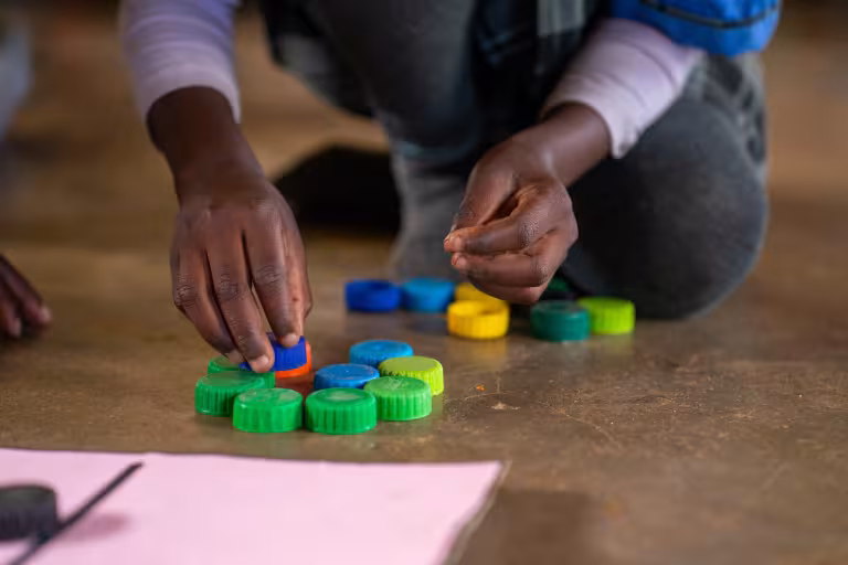A child’s hands arrange colourful plastic bottle caps on the floor during a classroom learning activity.