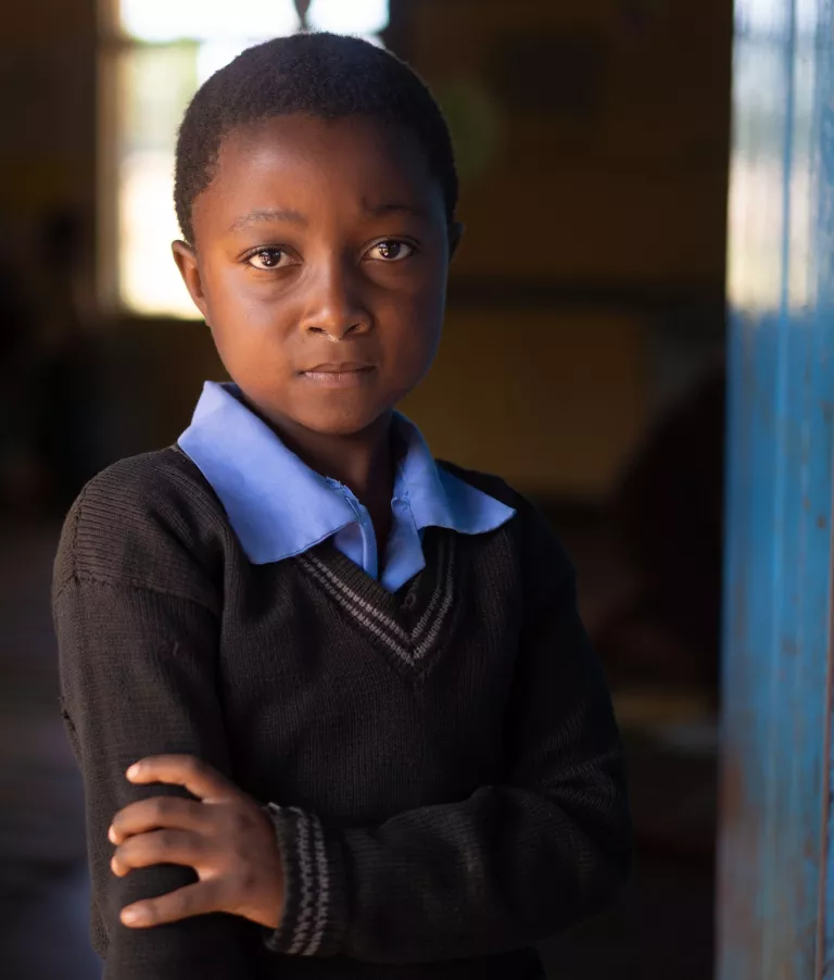 Blessings, a 6-year-old from Zambia is standing outside her classroom. She put one of her arms over the other looking directly into the camera lens.