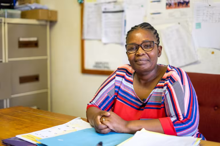 A woman in a colorful striped shirt sits confidently at a desk with papers, against a backdrop of a bulletin board, in a classroom setting.