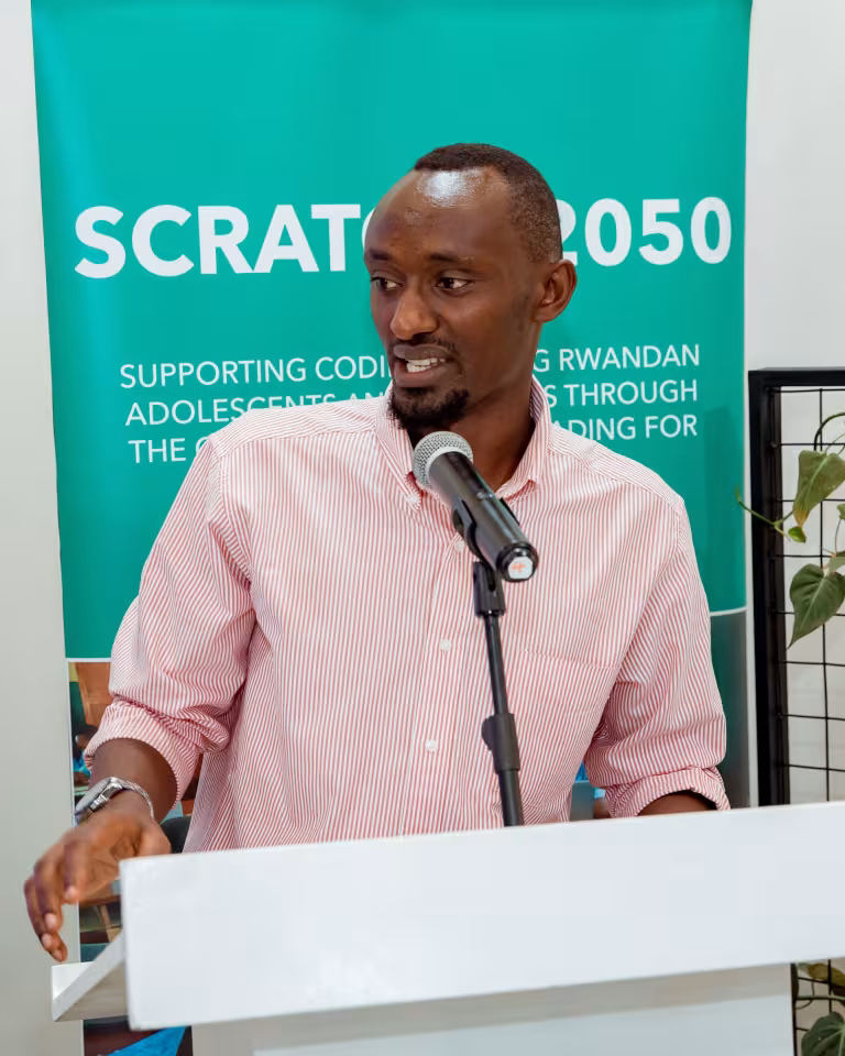 A man in a striped shirt speaks at a podium with a microphone. A banner behind him reads "SCRATCH2050," highlighting coding support for Rwandan adolescents. The atmosphere is professional and engaging.