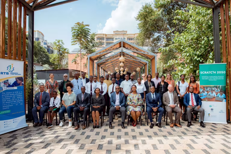 A diverse group of people, including men and women in formal attire, are posed in rows for a group photo under an outdoor structure with banners on each side. The setting is bright, with greenery and a building in the background, conveying a professional and collaborative tone.