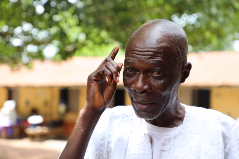 Close-up portrait of an elderly Black man outdoors, wearing a white traditional tunic, raising his index finger to his temple in a thoughtful gesture. The bald man has expressive facial features and a serious, contemplative expression. The background shows a blurred rural setting with green trees and a light-colored building, captured in natural daylight with shallow depth of field.
