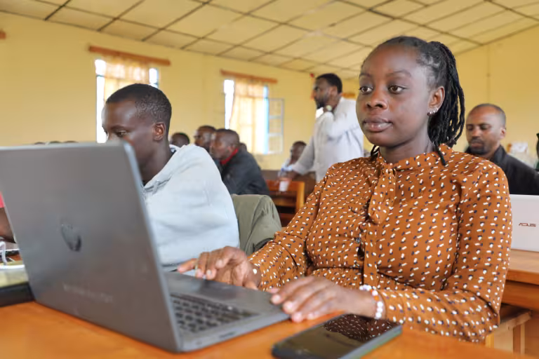 Young African woman using a laptop in a classroom computer lab, focused female student typing on notebook computer during digital skills training session, diverse group of adult learners in background, modern education environment with wooden desks and natural window light, technology in higher education, professional development and e-learning concept.