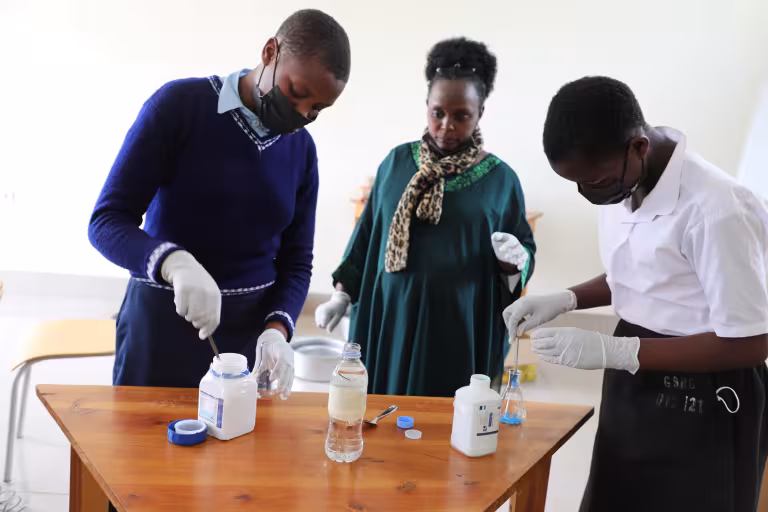 Two students and a teacher conduct a science experiment at a wooden table. They wear masks and gloves, focused on mixing chemicals in bottles.
