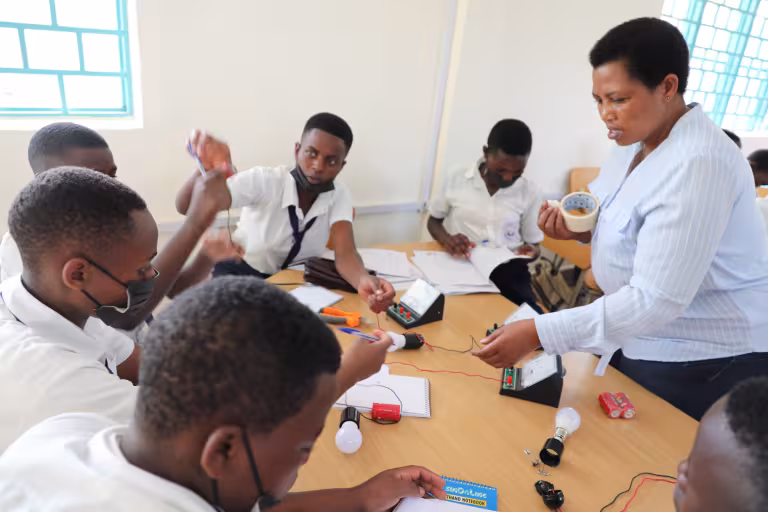 A teacher instructs attentive students on an electrical circuit project at a table. The classroom is bright, conveying an atmosphere of focused learning.