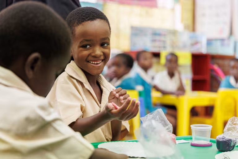 A smiling child shapes clay during a classroom learning activity while other learners work in the background.