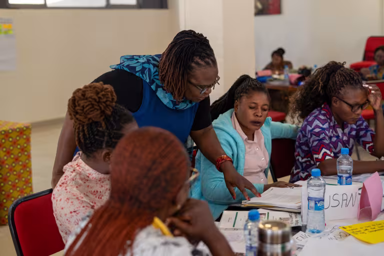 Women collaborate around a classroom table, reviewing papers and writing during a workshop; water bottles and colorful supplies fill the scene.